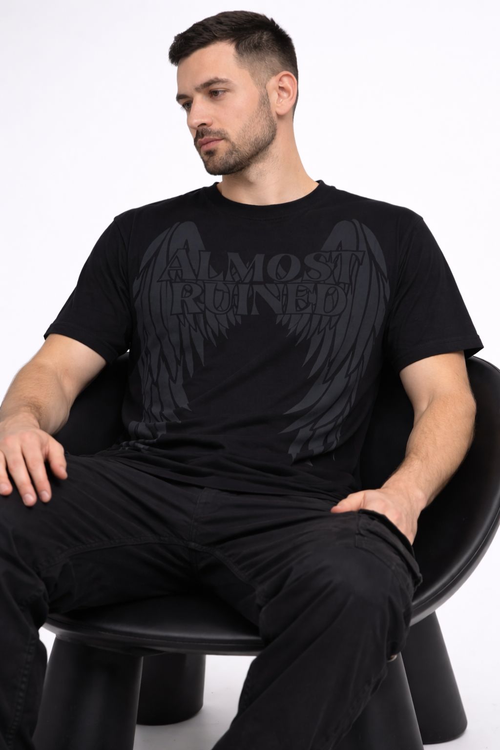 Man wearing a black t-shirt with a graphic design sitting on a chair against a white background