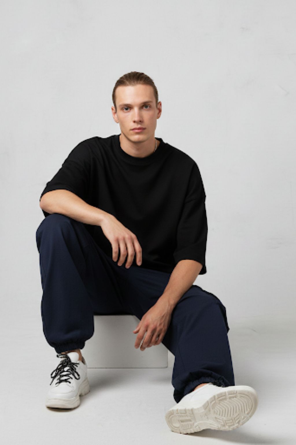 Man wearing a black sweater and navy pants sitting on a white chair against a light gray background